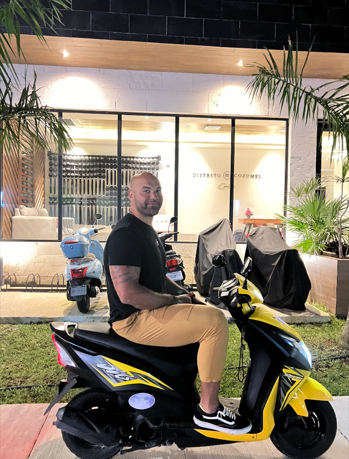 Screenshot_20251214-134209 – Jet Ski Cozumel Bearded man in a black shirt and tan pants sits on a yellow and black scooter in front of a glass storefront at night, with potted plants nearby.