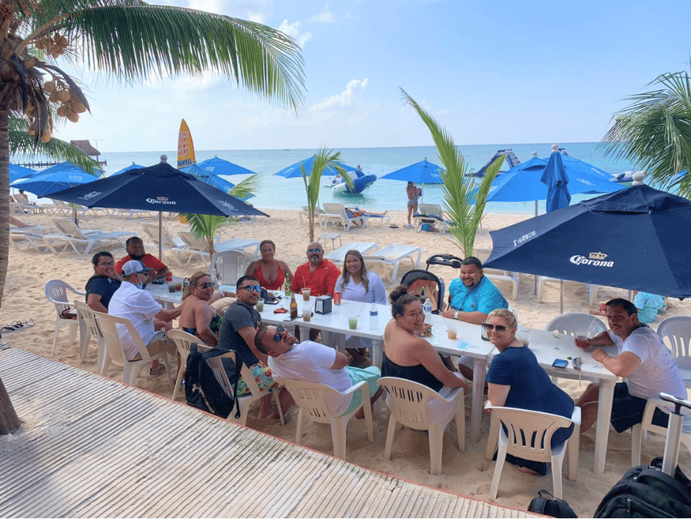 Group of people seated around a long white table on a sandy beach under blue umbrellas, with the ocean in the background.
