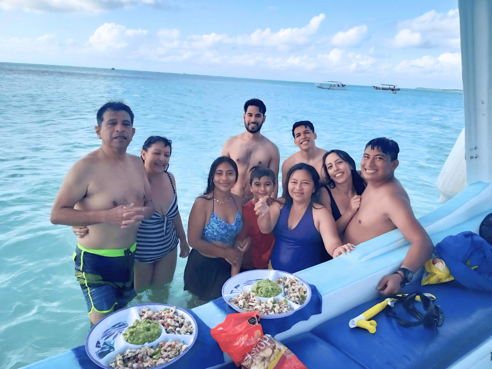 A Cozumel Private snorkel tour showing Nine people stand in the turquoise sea on a blue boat, smiling at the camera with two bowls of salad and sides in front of them on deck.